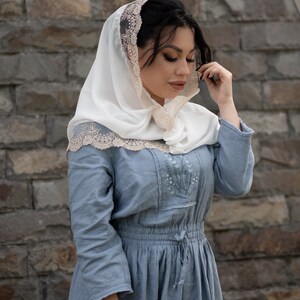 May include: A woman wearing a white head covering with a delicate lace trim and a light blue dress. The dress features embroidered floral details and a drawstring waist. The background is a textured stone wall.