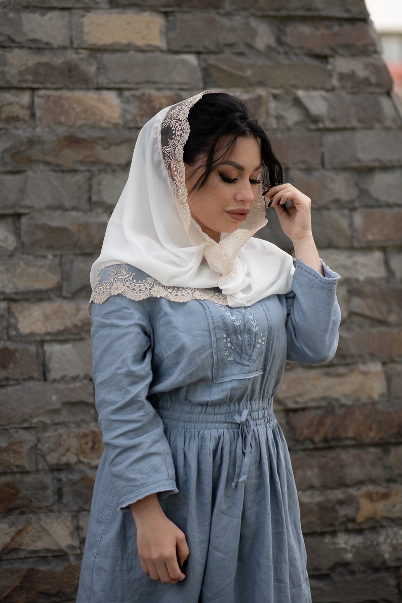 May include: A woman wearing a white head covering with a delicate lace trim and a light blue dress. The dress features embroidered floral details and a drawstring waist. The background is a textured stone wall.