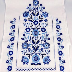 May include: A white rectangular table runner featuring intricate embroidery in shades of navy and light blue. The design displays a central vertical floral motif with symmetrical stems, leaves, and blossoms, flanked by two vertical floral borders.