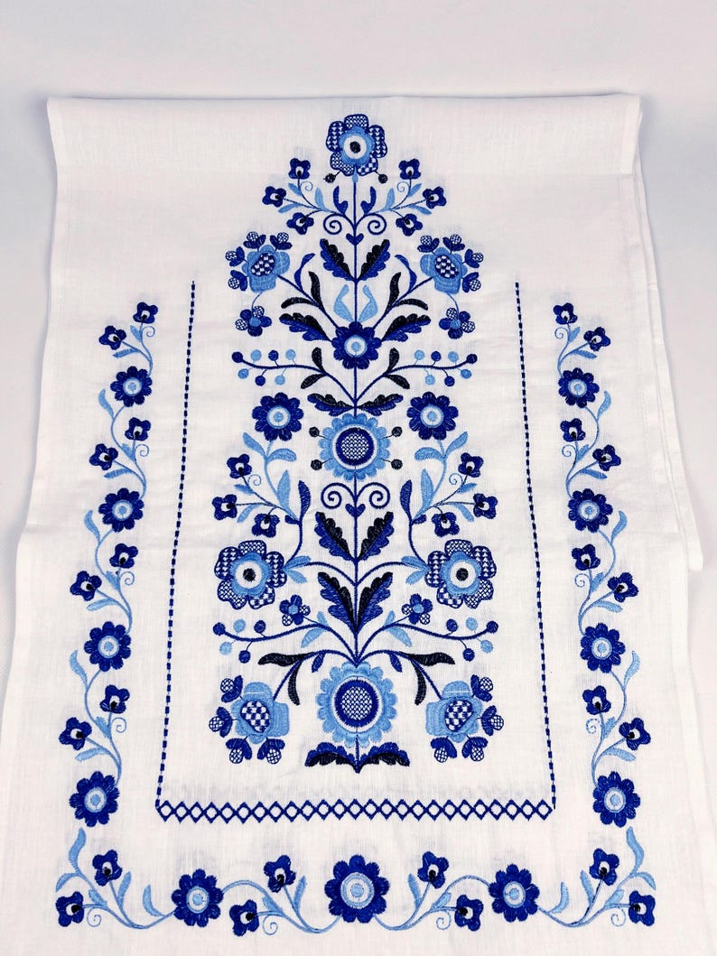 May include: A white rectangular table runner featuring intricate embroidery in shades of navy and light blue. The design displays a central vertical floral motif with symmetrical stems, leaves, and blossoms, flanked by two vertical floral borders.