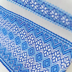 May include: Close-up of a white fabric with a detailed blue geometric pattern. The design features diamond and zigzag shapes in varying shades of blue, creating a textured and visually appealing surface. The fabric's intricate design suggests a decorative or textile item.