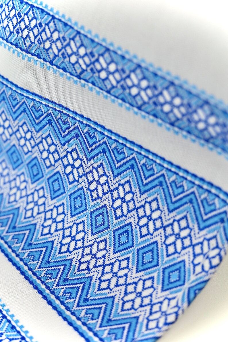 May include: Close-up of a white fabric with a detailed blue geometric pattern. The design features diamond and zigzag shapes in varying shades of blue, creating a textured and visually appealing surface. The fabric's intricate design suggests a decorative or textile item.