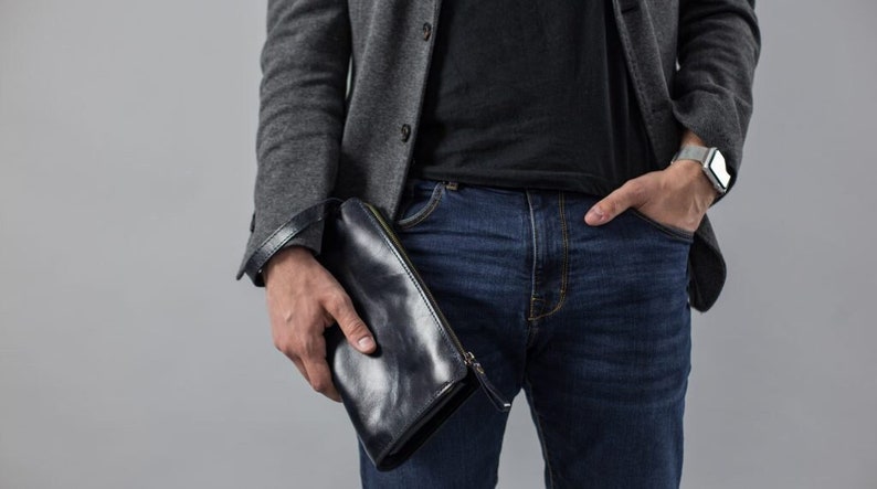 May include: A person wearing a gray blazer, black t-shirt, blue jeans, and a silver watch. They are holding a black leather clutch.