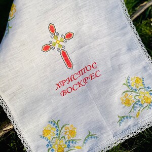 以下が含まれることがあります： 白いリネンの布に刺繍された十字架と花のアクセント。十字架は赤、緑、黄色で、「Христос Воскрес」の文字が赤色で書かれています。縁は白いレースでトリミングされ、花は黄色と青色です。