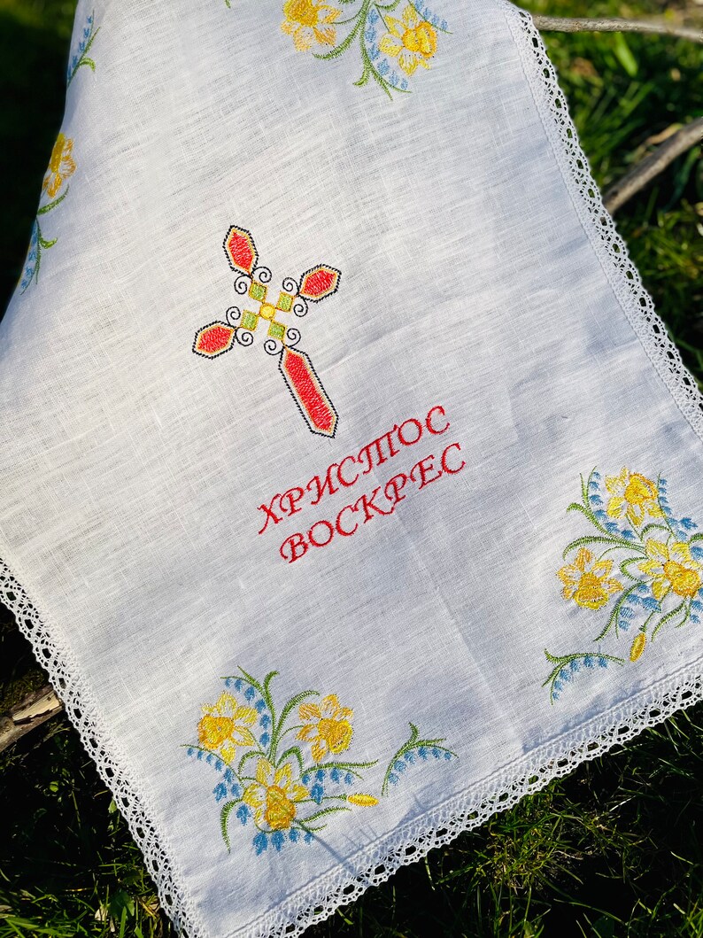 以下が含まれることがあります： 白いリネンの布に刺繍された十字架と花のアクセント。十字架は赤、緑、黄色で、「Христос Воскрес」の文字が赤色で書かれています。縁は白いレースでトリミングされ、花は黄色と青色です。