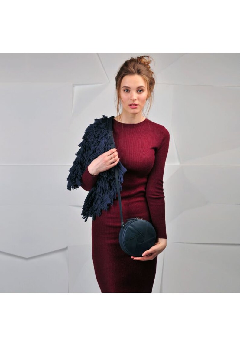 Puede incluir: Una mujer lleva un vestido granate y un c&aacute;rdigan azul con flecos y sostiene un bolso redondo de bandolera azul oscuro con un dise&ntilde;o geom&eacute;trico.