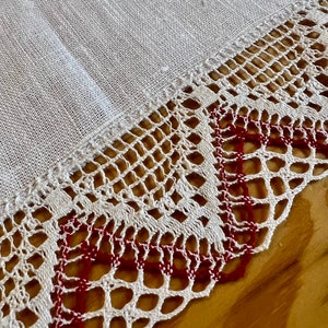 May include: White linen tablecloth with a delicate crocheted lace border. The lace has a red thread woven into the design, creating a subtle contrast.
