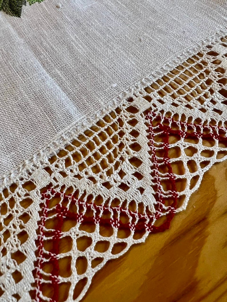 May include: White linen tablecloth with a delicate crocheted lace border. The lace has a red thread woven into the design, creating a subtle contrast.