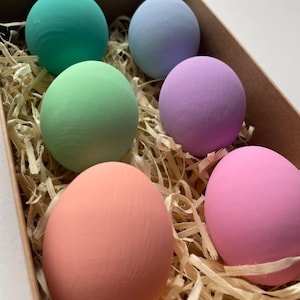 May include: Six painted eggs in pastel shades of green, blue, purple, pink, and peach, presented in a cardboard box with straw. The eggs have a smooth texture and are a solid colour. A decorative Easter theme.