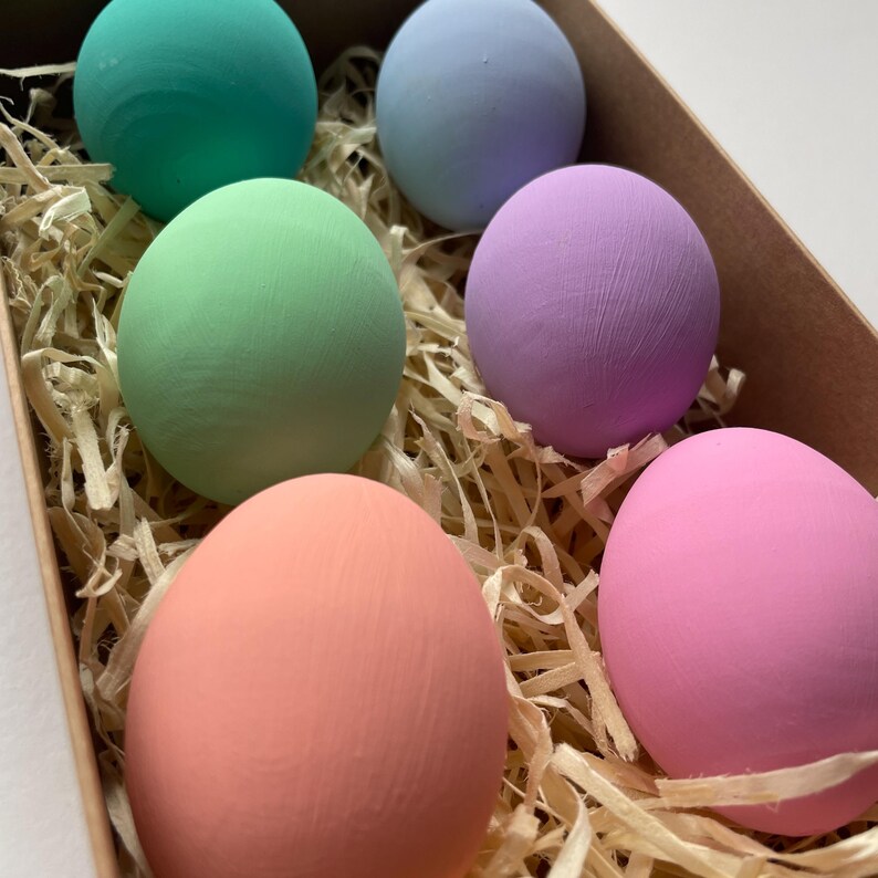 May include: Six painted eggs in pastel shades of green, blue, purple, pink, and peach, presented in a cardboard box with straw. The eggs have a smooth texture and are a solid colour. A decorative Easter theme.