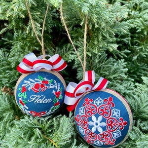 K&ouml;nnte beinhalten: Zwei blaue und wei&szlig;e Weihnachtskugeln mit rot-wei&szlig;em Band. Eine Kugel hat einen Kranz mit Stechpalme und Glocken und den Namen "Helen" bestickt. Die andere Kugel hat ein Schneeflockenmuster.