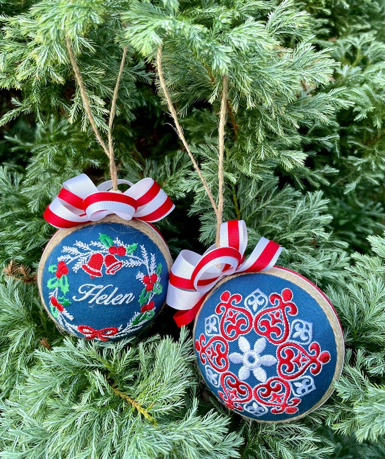 K&ouml;nnte beinhalten: Zwei blaue und wei&szlig;e Weihnachtskugeln mit rot-wei&szlig;em Band. Eine Kugel hat einen Kranz mit Stechpalme und Glocken und den Namen "Helen" bestickt. Die andere Kugel hat ein Schneeflockenmuster.