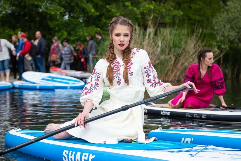 May include: A woman in a white dress with red and blue floral embroidery sits on a blue paddleboard, holding a paddle. Another woman in a pink dress is in the background. The paddleboard has the word "SHARK" on it.