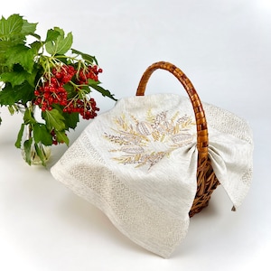 May include: A wicker basket with a curved handle, covered by a beige linen cloth. The cloth has embroidered floral designs in gold and brown. A vase of red berries and green leaves is to the left.