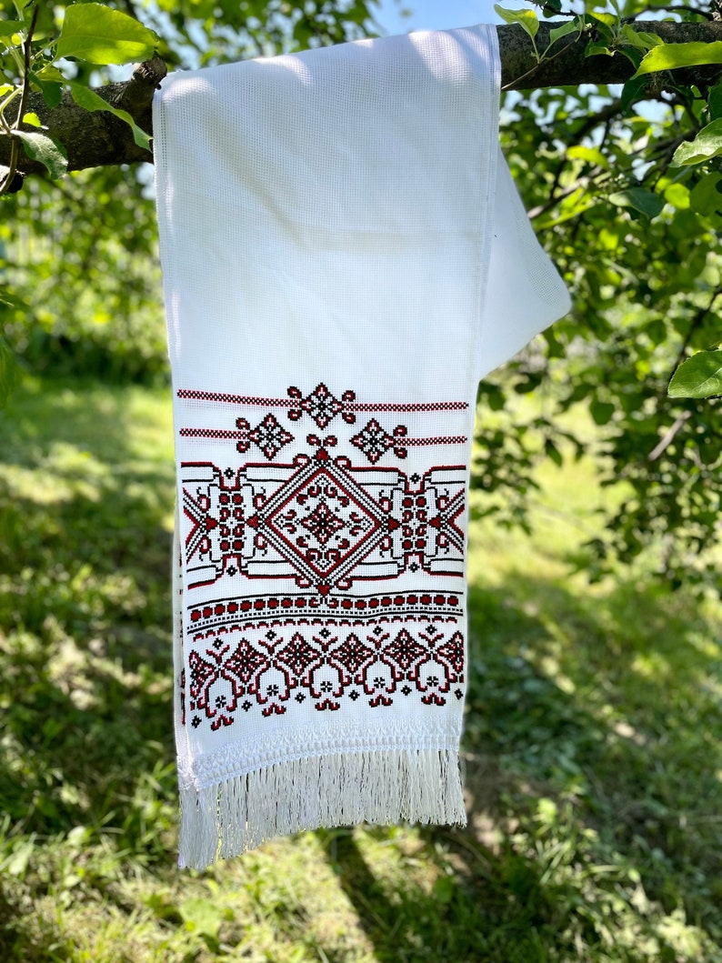 Puede incluir: Toalla blanca bordada con un detallado patr&oacute;n geom&eacute;trico en rojo y negro. La toalla cuelga de una rama de &aacute;rbol, mostrando una franja en la parte inferior. El dise&ntilde;o presenta una forma de diamante central y motivos florales.