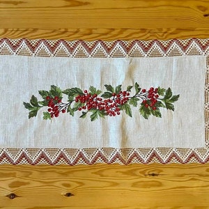 May include: White linen table runner with a red and green embroidered floral design. The runner has a delicate lace border.