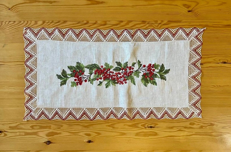 May include: White linen table runner with a red and green embroidered floral design. The runner has a delicate lace border.