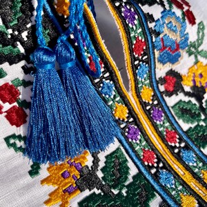 May include: Close-up of a white embroidered shirt with blue tassels. The embroidery features a colorful floral pattern with black, red, yellow, green, and blue thread.