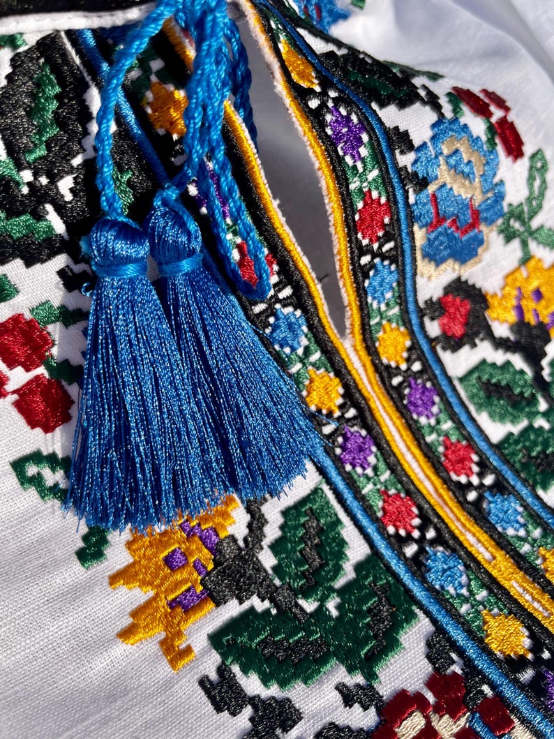 May include: Close-up of a white embroidered shirt with blue tassels. The embroidery features a colorful floral pattern with black, red, yellow, green, and blue thread.