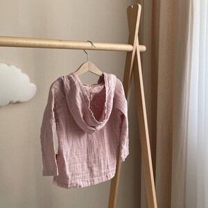 May include: A pale pink hooded sweatshirt with a textured, gauze-like fabric. The sweatshirt is hanging on a wooden hanger.