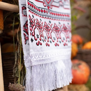 Puede incluir: Textil blanco bordado con patrones geom&eacute;tricos rojos y negros y flecos. El textil est&aacute; colgado de una estructura de madera, con un fondo borroso con calabazas y follaje.