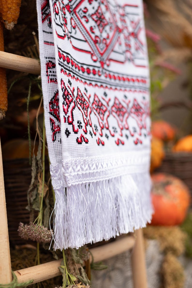Puede incluir: Textil blanco bordado con patrones geom&eacute;tricos rojos y negros y flecos. El textil est&aacute; colgado de una estructura de madera, con un fondo borroso con calabazas y follaje.