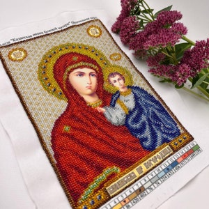May include: A beaded religious icon depicting the Virgin Mary and Child, with a gold halo and red and blue robes. The artwork is on a white fabric background, with text in the upper left corner and a color chart at the bottom. Purple flowers are in the background.