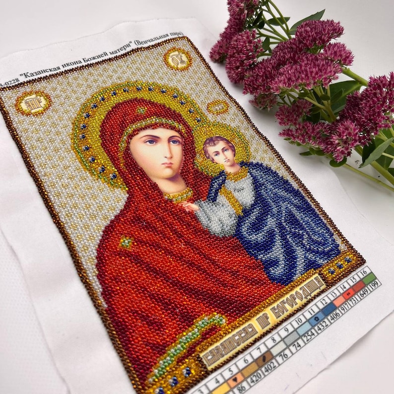 May include: A beaded religious icon depicting the Virgin Mary and Child, with a gold halo and red and blue robes. The artwork is on a white fabric background, with text in the upper left corner and a color chart at the bottom. Purple flowers are in the background.