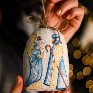 a handmade linen Christmas ornament embroidered with a Nativity scene showing Mary, Joseph, and baby Jesus in blue and gold thread, decorated with a jute bow
