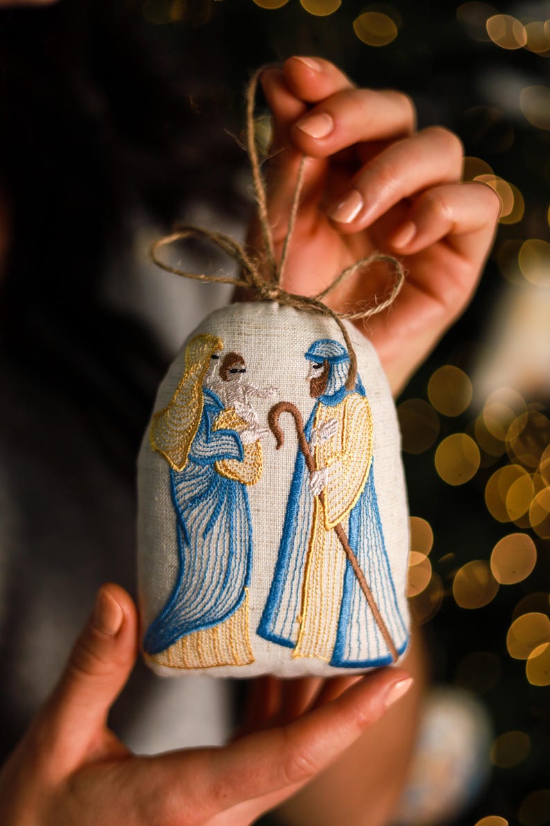 a handmade linen Christmas ornament embroidered with a Nativity scene showing Mary, Joseph, and baby Jesus in blue and gold thread, decorated with a jute bow
