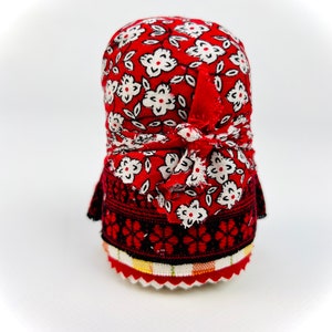 Op de afbeelding: Een traditionele Russische nestpop, of matrjosjkapop, met een rood en wit bloemenpatroon. De pop draagt een rode hoofddoek en een zwarte en rode geborduurde rok.