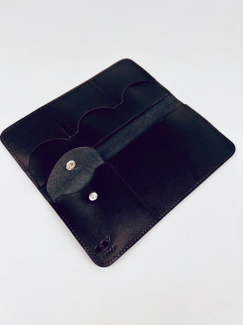 K&ouml;nnte beinhalten: Eine schwarze Ledergeldb&ouml;rse mit mehreren Kartenf&auml;chern und einem Druckknopfverschluss. Die Geldb&ouml;rse hat ein kleines Logo, das in die rechte untere Ecke gepr&auml;gt ist.