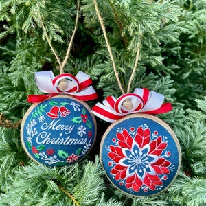 K&ouml;nnte beinhalten: Zwei handgemachte Weihnachtskugeln aus blauem Stoff mit roten und wei&szlig;en Stickereien. Eine Kugel hat den Text "Frohe Weihnachten" und die andere ein Blumenmuster.