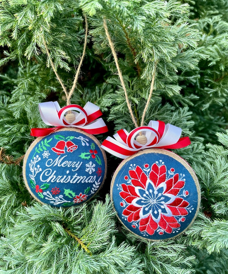 K&ouml;nnte beinhalten: Zwei handgemachte Weihnachtskugeln aus blauem Stoff mit roten und wei&szlig;en Stickereien. Eine Kugel hat den Text "Frohe Weihnachten" und die andere ein Blumenmuster.