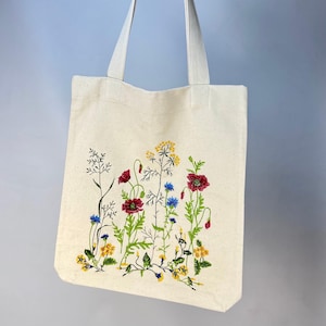 以下が含まれることがあります： クリーム色のトートバッグ。野花が刺繍されています。赤いポピー、黄色と青の花、緑の茎のデザインです。バッグにはクリーム色のハンドルが2つ付いています。