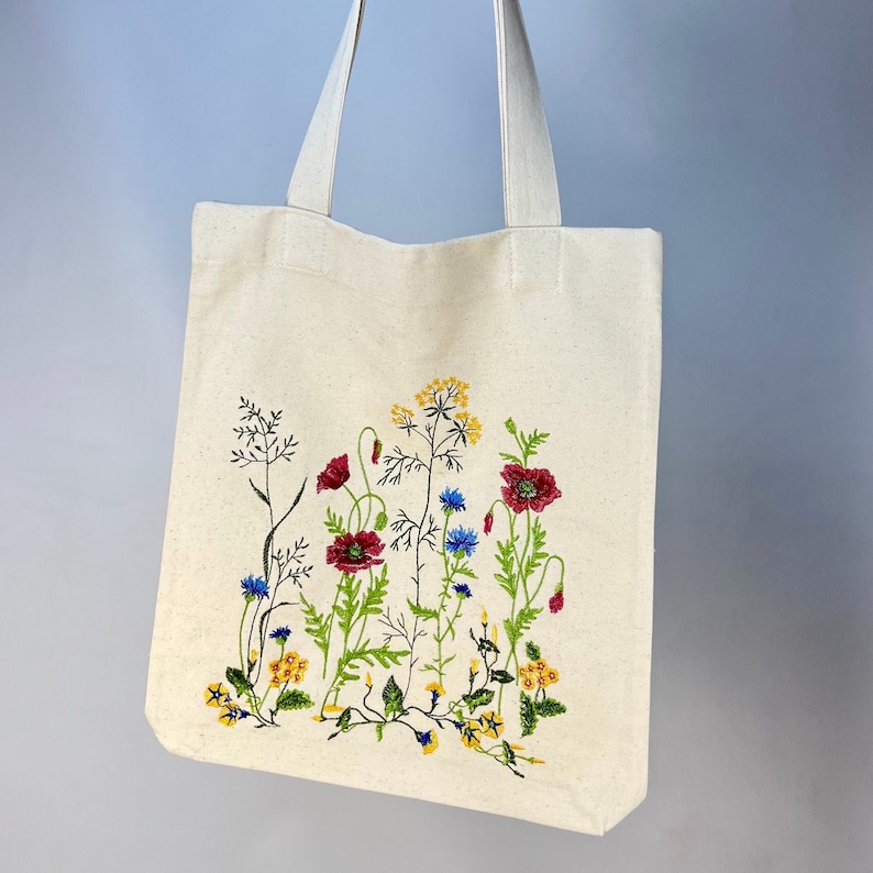 以下が含まれることがあります： クリーム色のトートバッグ。野花が刺繍されています。赤いポピー、黄色と青の花、緑の茎のデザインです。バッグにはクリーム色のハンドルが2つ付いています。