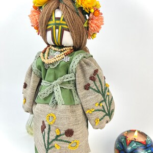 Puede incluir: Mu&ntilde;eca hecha a mano con corona floral y vestimenta tradicional. La mu&ntilde;eca tiene una cara blanca con un dise&ntilde;o en forma de cruz, vistiendo un atuendo verde y beige con bordados florales. Tambi&eacute;n se ven un collar de cuentas y una vela decorativa.