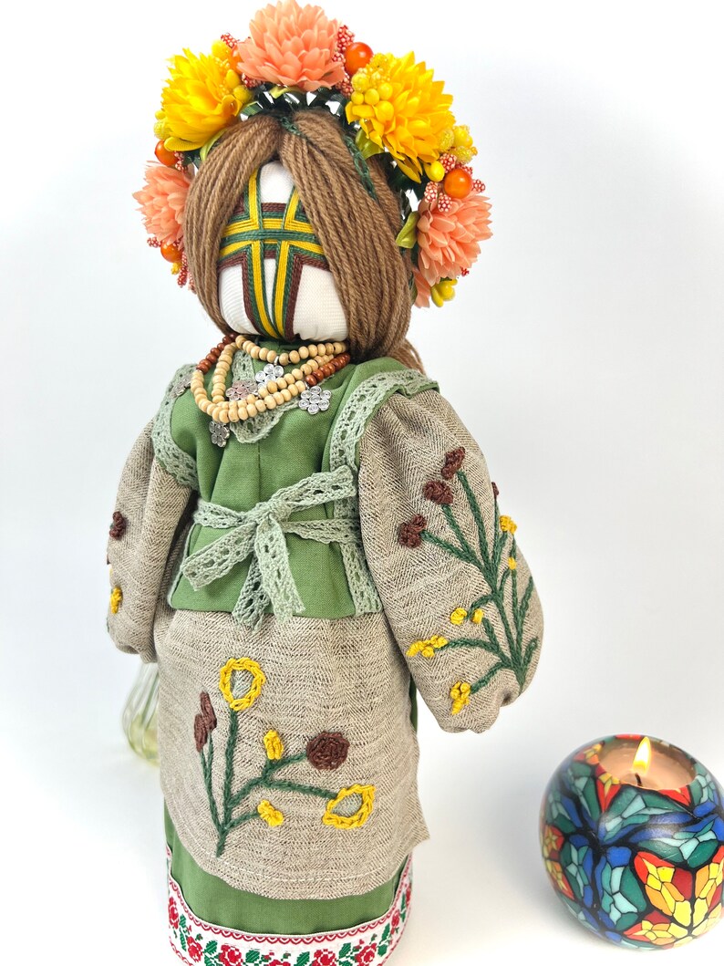 Puede incluir: Mu&ntilde;eca hecha a mano con corona floral y vestimenta tradicional. La mu&ntilde;eca tiene una cara blanca con un dise&ntilde;o en forma de cruz, vistiendo un atuendo verde y beige con bordados florales. Tambi&eacute;n se ven un collar de cuentas y una vela decorativa.