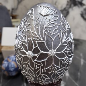 Puede incluir: Un huevo decorativo plateado con dise&ntilde;os florales blancos. El huevo presenta contornos blancos detallados de flores, hojas y peque&ntilde;os puntos, creando una apariencia texturizada y en relieve. Un art&iacute;culo &uacute;nico y artesanal.