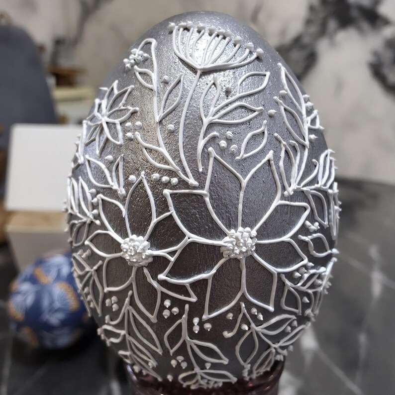 Puede incluir: Un huevo decorativo plateado con dise&ntilde;os florales blancos. El huevo presenta contornos blancos detallados de flores, hojas y peque&ntilde;os puntos, creando una apariencia texturizada y en relieve. Un art&iacute;culo &uacute;nico y artesanal.