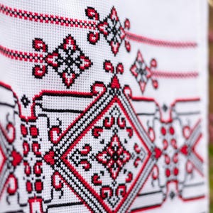 Puede incluir: Primer plano de tela blanca con intrincado bordado de punto de cruz. El dise&ntilde;o presenta patrones geom&eacute;tricos en rojo y negro, incluyendo diamantes y motivos florales. La labor de aguja detallada crea una superficie texturizada.