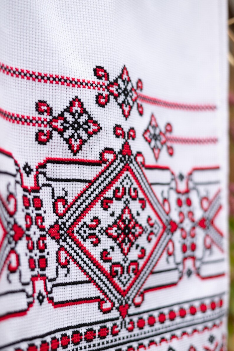 Puede incluir: Primer plano de tela blanca con intrincado bordado de punto de cruz. El dise&ntilde;o presenta patrones geom&eacute;tricos en rojo y negro, incluyendo diamantes y motivos florales. La labor de aguja detallada crea una superficie texturizada.