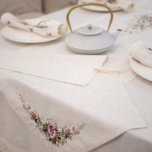 May include: A white linen tablecloth with embroidered floral designs. The tablecloth is set with a white teapot with a gold handle, two white plates, and two white napkins.