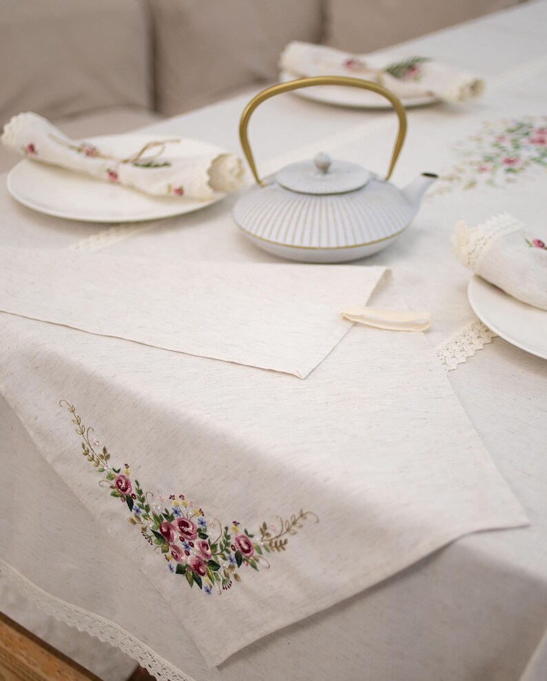 May include: A white linen tablecloth with embroidered floral designs. The tablecloth is set with a white teapot with a gold handle, two white plates, and two white napkins.