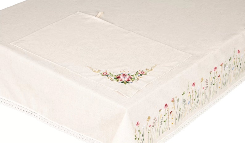 May include: Cream-colored linen tablecloth with embroidered floral details. A matching placemat is placed on the table. The edge of the tablecloth features a delicate floral border with red, pink, and yellow flowers.