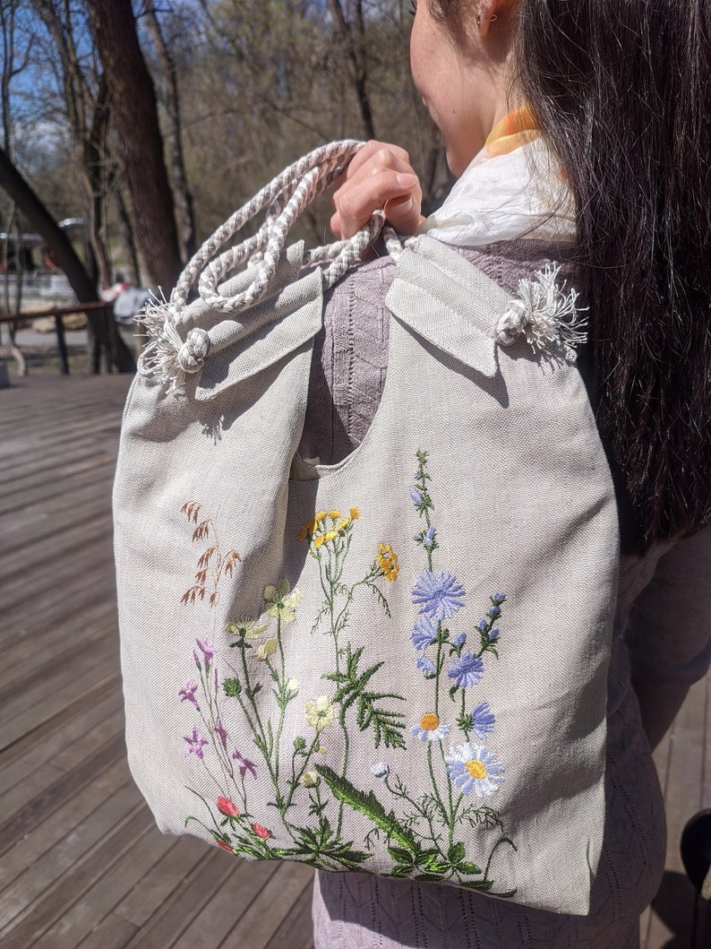 Op de afbeelding: Een beige canvas draagtas met een wit touw handvat en geborduurde wilde bloemen in tinten van blauw, geel, groen en roze.