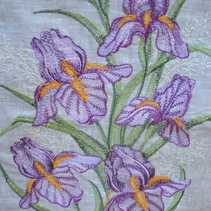 May include: Embroidered purple iris flowers with yellow centres on a beige linen fabric background.
