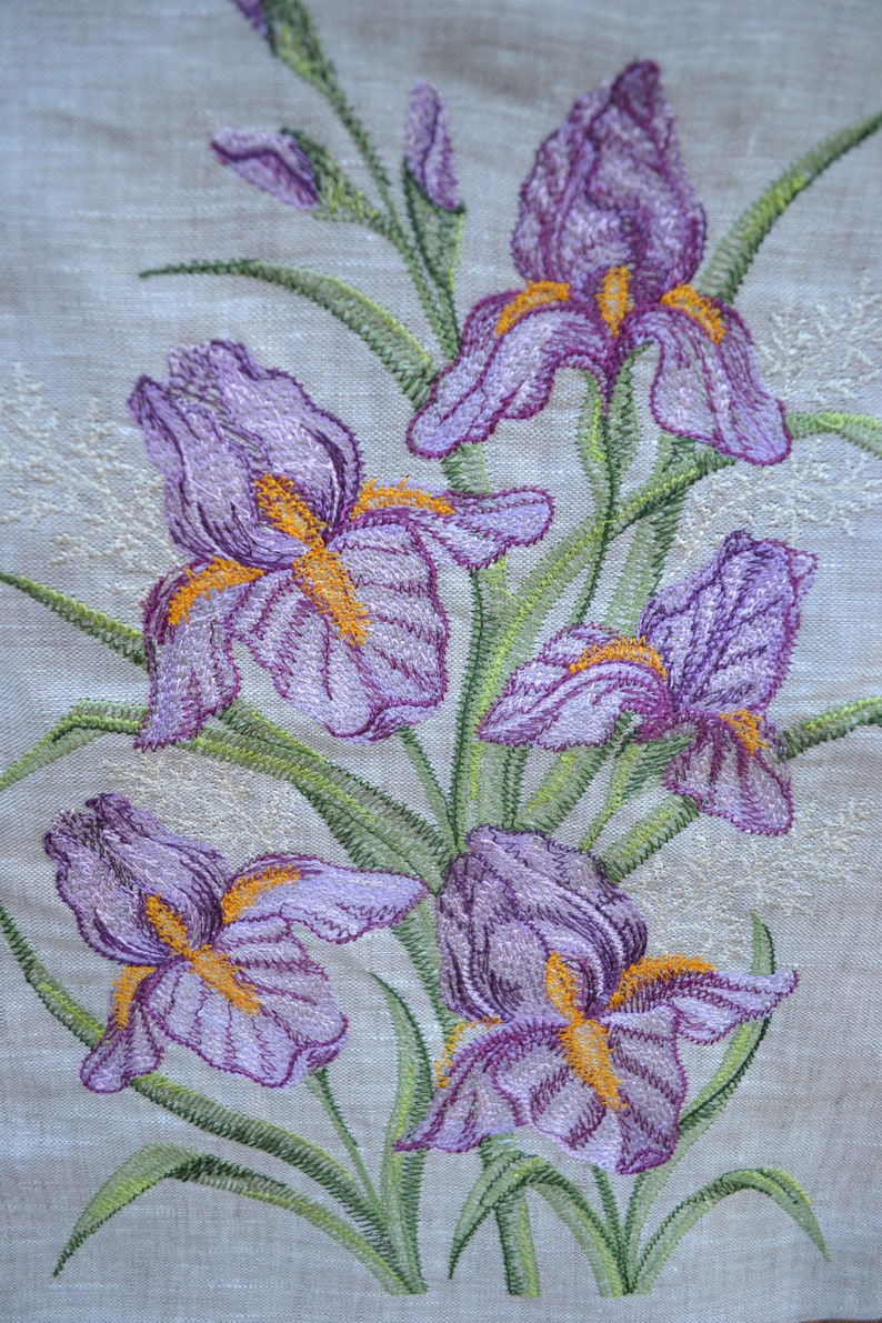 May include: Embroidered purple iris flowers with yellow centres on a beige linen fabric background.