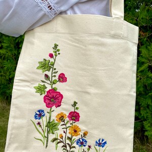 以下が含まれることがあります： 白いキャンバス地のトートバッグに、花の刺繍が施されています。刺繍には、ピンク、黄色、青の花と緑の葉が描かれています。