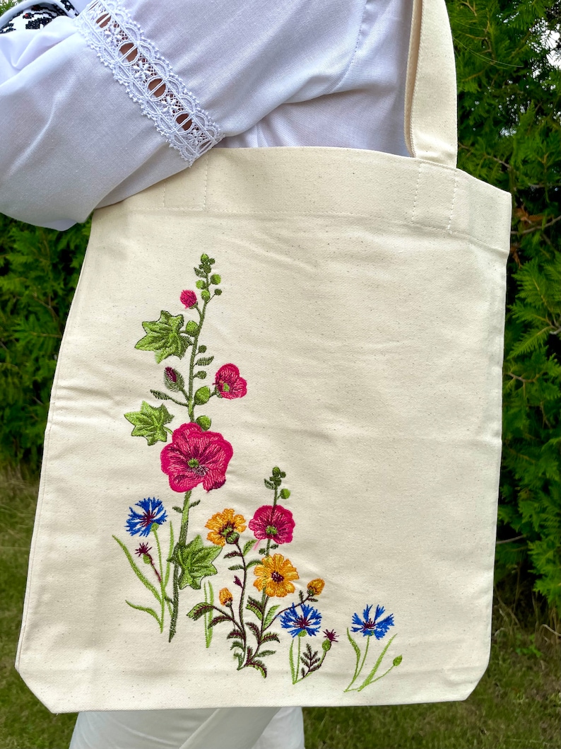 以下が含まれることがあります： 白いキャンバス地のトートバッグに、花の刺繍が施されています。刺繍には、ピンク、黄色、青の花と緑の葉が描かれています。